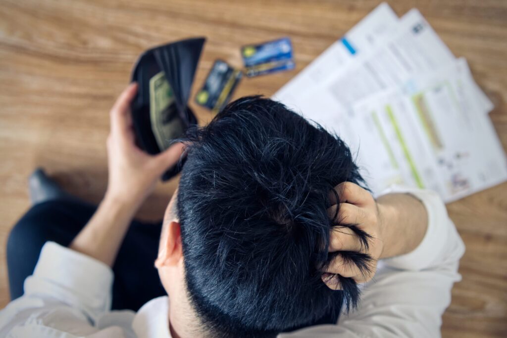 Top view of stressed young Asian man holding wallet with money and thinking about finding money to pay credit card debt and all bills. He is holding head by another hand. Financial problem concept.